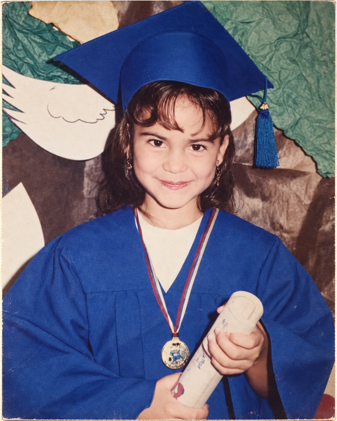 Genesis Perera de niña en su graduación, con toga y birrete azules, una medalla y su diploma.